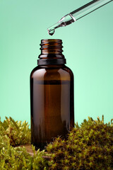 Glass pipette with drop of essential oil over brown bottle among the moss against green background. Essence liquid for skin care or alternative medicine.