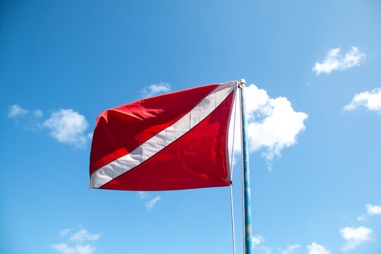 A red and white scuba diving flag flapping in the wind, isolated, with a diagonal stripe attached to flag pole against bright blue sky with clouds, Barbados, Caribbean. Sport, recreation symbol.