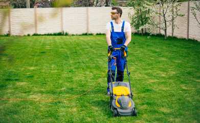 Young man mows the lawn using an electric lawn mower in a special worker suit near a large country house in the backyard
