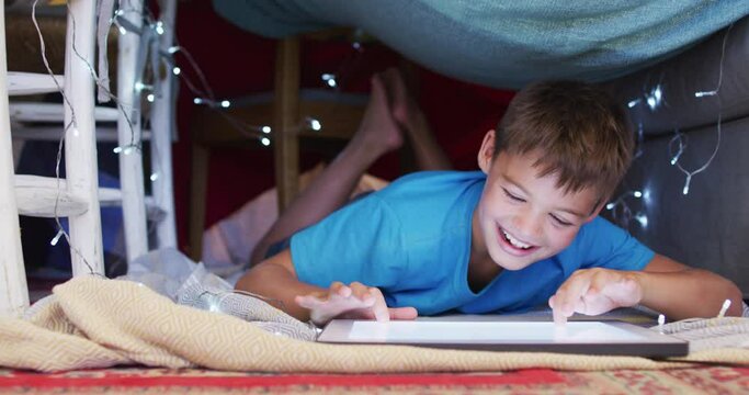 Caucasian Boy Smiling While Using Digital Tablet Under The Blanket Fort At Home