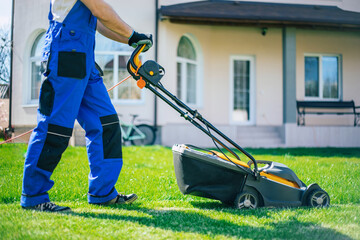 Young man mows the lawn using an electric lawn mower in a special worker suit near a large country house in the backyard - obrazy, fototapety, plakaty