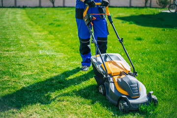 Young man mows the lawn using an electric lawn mower in a special worker suit near a large country...