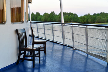 On the deck of the ship, there are chairs near the windows of the cabins. View from the deck to the green shore.
