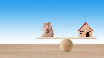 Model house on wooden seesaw balancing with money bag on blue background. Property investment and...