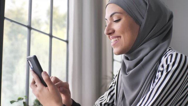 pleased muslim arab girl wearing hijab headscarf using smart phone mobile at the cafe',young middle easter woman sits by the window holding smartphone enjoying a drink