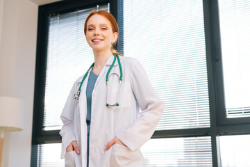 Portrait of positive female doctor in white coat standing on background of window in sunny day in light medical clinic office. Young redhead woman physician posing with stethoscope looking at camera.