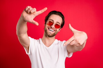 Fototapeta premium Photo of cheerful positive young man wear sunglass make fingers frame camera isolated on red color background