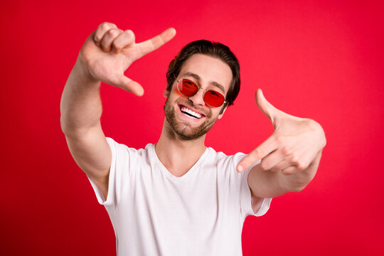Photo of cheerful positive young man wear sunglass make fingers frame camera isolated on red color background