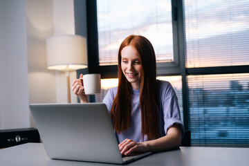 Happy young woman using laptop computer and drinking coffee at home workplace. Close-up of business woman watching news at laptop screen in creative office. Female professional working notebook.