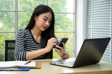 Happy young Asian woman smiling writing memo on pocket PC taking data from laptop in living room. teenage work from home concept