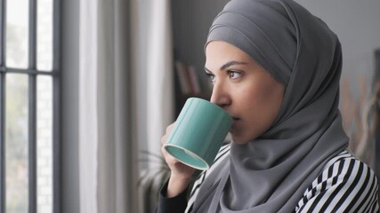 pensive muslim arab business woman wearing hijab headscarf standing by the window enjoying a coffee cup,close up portrait of thoughtful middle eastern woman drinking and looking outside - Powered by Adobe