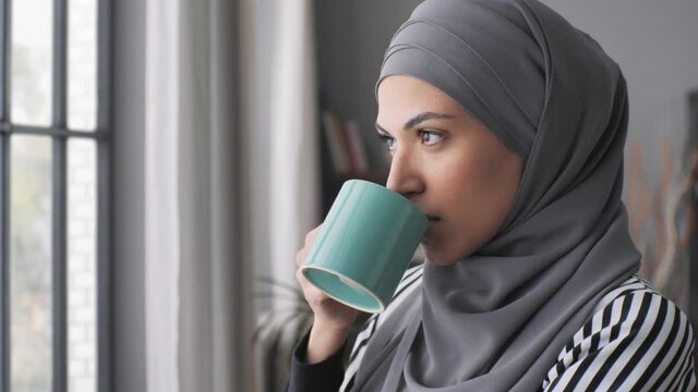 pensive muslim arab business woman wearing hijab headscarf standing by the window enjoying a coffee cup,close up portrait of thoughtful middle eastern woman drinking and looking outside