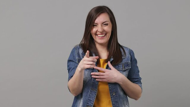Excited cheerful fun brunette young woman 20s years old in denim jacket yellow t-shirt look camera laugh smiling watch comedy movie point index finger on you isolated on grey color background studio