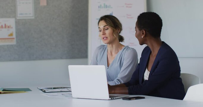 Two diverse female colleagues sitting at desk with laptop and discussing in office