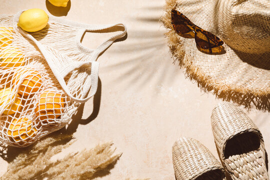 Summer flat lay on beige background. Straw hat, sunglasses, slippers and lemons in eco friendly mesh shopping bag. Palm shadow and sunlight, sun. Minimal summer travel fashion concept. 
