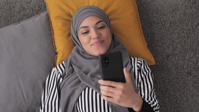 muslim girl with headscarf hijab lying on the floor holding smart phone makes a video call conference,young middle eastern woman having an online videocall with friend or family