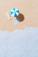 White and blue beach umbrella, sun chair and blue and white striped towel on sand and paper waves. Beach set for sunny days. Summer holiday concept. top view with copy space. Vertical image.