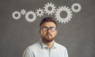 Creative thinking and ideas. Young businessman thinking about project optimization standing on a gray background. Serious millennial man with glasses looks thoughtfully up at the gears above his head.