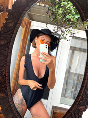 Pretty woman at home take photo selfie in mirror on mobile phone for stories and posts in social media, vertical frame, wearing black summer swimsuit, large tattoo on leg, fit and tanned