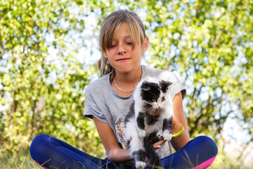 Naklejka na ściany i meble Defocus blonde little girl holding and caress cat, black and white small beautiful kitten. Nature green summer background. Girl holding and stroking pet. Love animals concept. Out of focus
