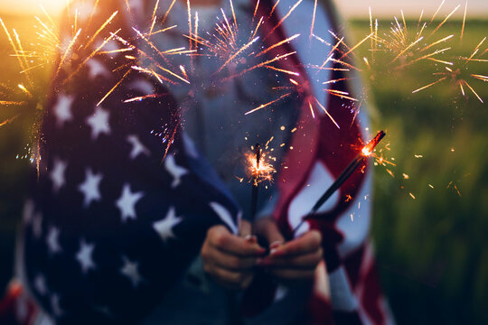 America celebrate 4th of July. Independence Day. Young woman holding bengal fire with American flag at sunset. 