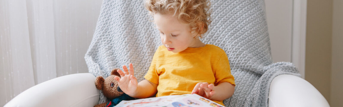 Cute baby boy toddler sitting at kids room and reading book. Early age kid child development literacy alphabet education. Candid home authentic childhood lifestyle. Web banner header.