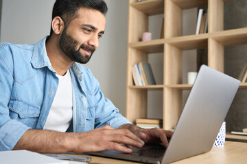 Handsome young Indian Hispanic businessman using computer working remotely from modern home office. Latin student having virtual training on laptop typing watching online webinar during quarantine.