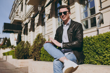 Young fashionable happy trendy fun smiling caucasian happy man 20s wear black leather jacket eyeglasses sitting near bulding and bushes resting in city downtown outdoors Concept of urban lifestyle