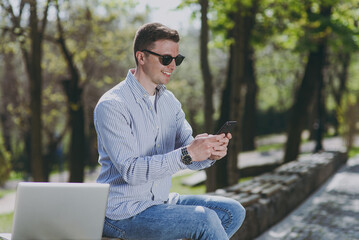 Side view young smiling fun happy caucasian freelancer man in shirt glasses sit on park bench work by laptop pc computer outdoors, using mobile cell phone wi-fi, look aside Concept of urban lifestyle.