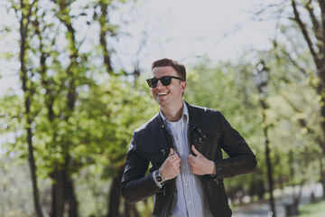 Young trendy stylish happy fun smiling caucasian man 20s wearing black leather jacket jeans eyeglasses strolling walking in green park looking aside. Concept of outdoors urban lifestyle spring season.