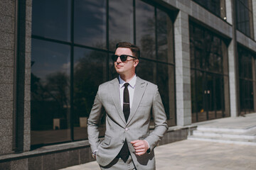 Young happy confident european successful employee business man 20s wear grey suit walking go near office glass wall building outdoors in downtown city center look aside. Achievement career concept.