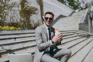 Young happy smiling businessman man 20s wear grey formal suit work by laptop pc computer browsing internet sitting on concrete steps in city center eat croissant have break lunch breakfast outdoors.