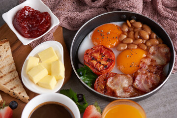 Traditional full English breakfast in cooking pan with fried eggs, bacon, beans, grilled tomatoes. toast, butter, jam on a wooden board, coffee and orange juice on stone background. top view.