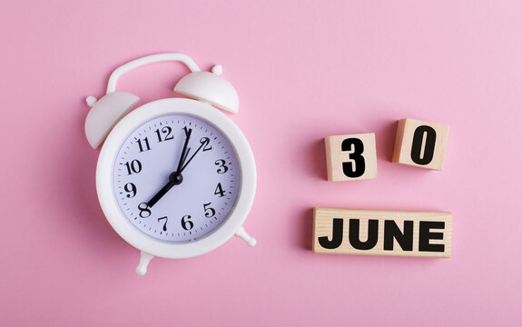 On a pink background, a white alarm clock and wooden cubes with the date of JUNE 30