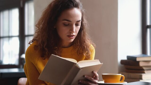 A focused pretty woman is reading a book sitting in the cafe inside
