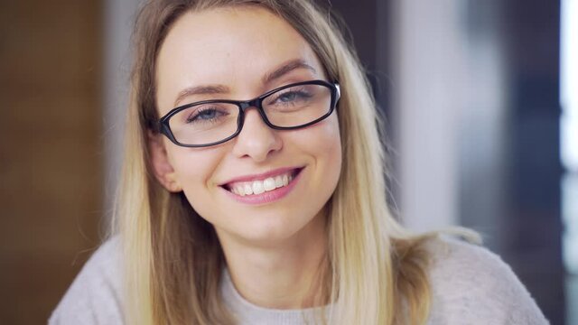 Close-up portrait a caucasian young blonde woman indoors looking at camera and smiling. Cute Happy Female with glasses in the office or at home with makeup and a beautiful smile and blue eyes. Closeup