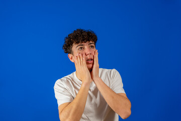 Attractive young man with curly hair with hands on face with scared gesture isolated on blue studio background