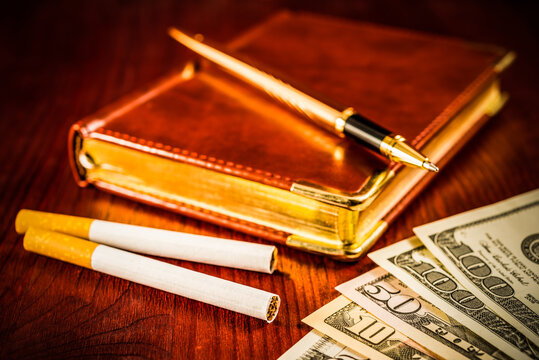 Money with a leather diary and cigarettes with golden pen on a mahogany table. Focus on the cigarettes, shallow deep of field