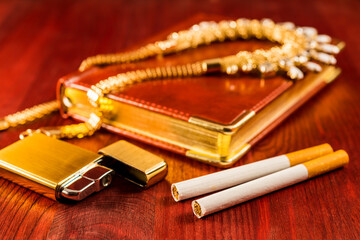 Golden lighter with a leather diary and cigarettes with jewellery on a mahogany table. Focus on the cigarettes