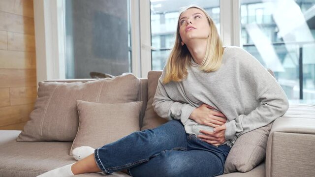 young woman sitting or lying on sofa or couch at home with severe abdominal pain. Female on bed holds her stomach with premenstrual, menstrual or poisoning. blonde girl suffers from abdominal cramps