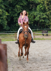Young woman wearing shirt riding brown horse in sand paddock by wooden fence, hair moving in air because of speed
