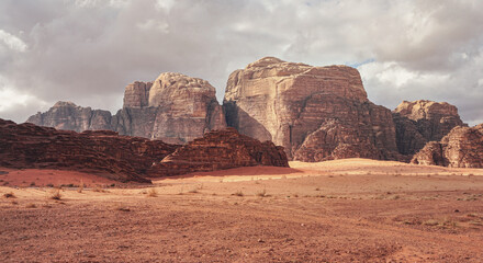 Rocky massifs on red sand desert, few dry grass cluster ground, cloudy sky in background, typical scenery in Wadi Rum, Jordan