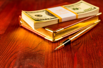 Pack of dollars on a leather diary with golden pen on a mahogany table. Focus on the golden pen