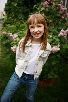smiling girl in a white denim jacket near a bush of blooming lilacs