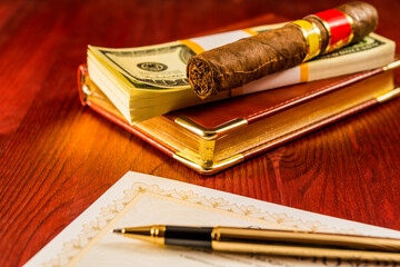 Pack of dollars and cuban cigar on the leather diary with bank check and golden pen on the table. Focus on the cuban cigar