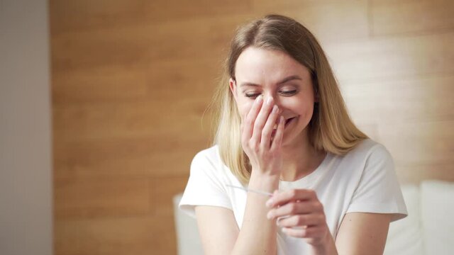 Happy caucasian young woman rejoices in positive pregnancy kit test for motherhood. Close up portrait of female at home checking result and emotions excited sitting on bed in bedroom. Happiness