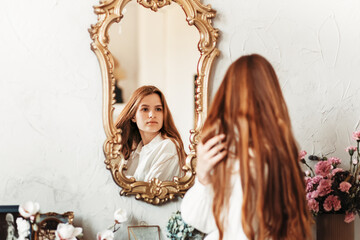 Young beautiful girl with long hair stands near the mirror in the morning in the bedroom