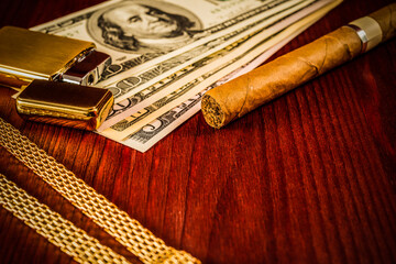 Cuban cigar and a gold chain with money and golden lighter on a table in mahogany. Focus on the...