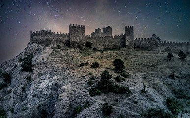 Medieval castle on a high rock at night. Stars on the sky. Medieval Genoese fortress in Crimea, Russia. Starry night in summer. Shrub growing on rocks. Ancient Fortress in Sudak. - obrazy, fototapety, plakaty