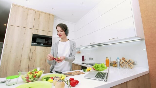 Sliding view of young attractive woman cooking salad, cutting fresh vegetables abd listenning to music in the headphones and dancing